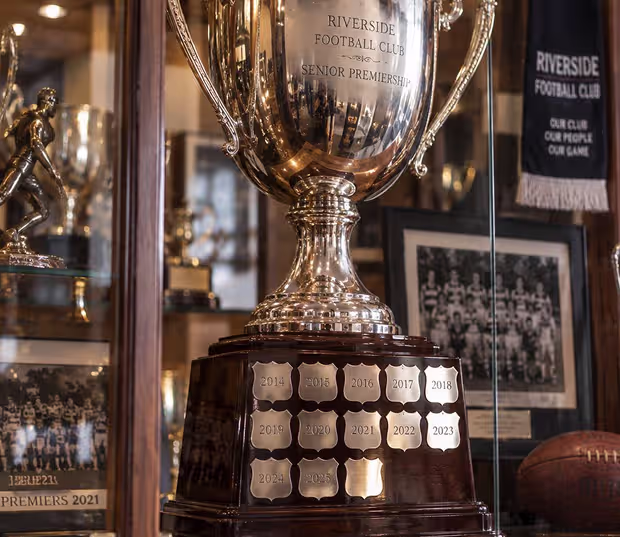 A perpetual premiership cup in a club cabinet, its base ringed with engraved year plates from 2014 onward and one blank plate still waiting.