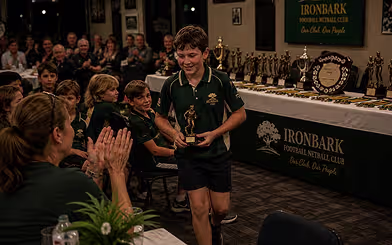 A junior player walking back to his seat holding his trophy while the other juniors and the room applaud.